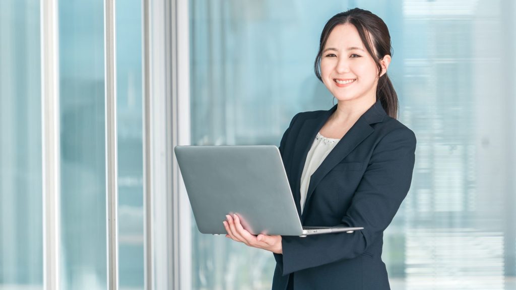オフィスの窓際で、ノートパソコンを抱えてこちらに向かって微笑む、紺色スーツの女性。